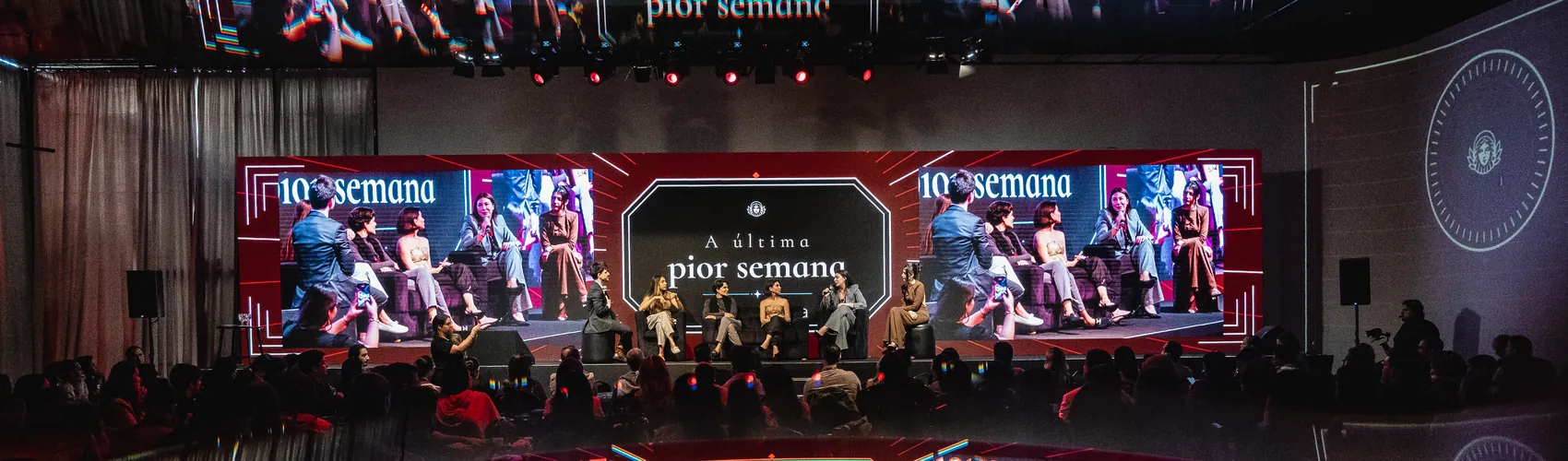 Três telões panorâmicos do evento presencial da Reconecta, com a plateia em silhueta